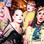 Four performers in bright drag posing together at the bar in a London queer venue