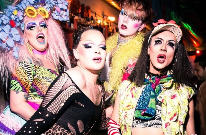 Four performers in bright drag posing together at the bar in a London queer venue