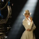 Jennifer Lopez in a white suit and fedora, lit by a shaft of light beside a vintage film camera on a soundstage, in a production still from Kiss of the Spider Woman