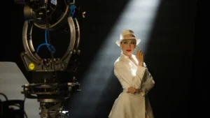 Jennifer Lopez in a white suit and fedora, lit by a shaft of light beside a vintage film camera on a soundstage, in a production still from Kiss of the Spider Woman