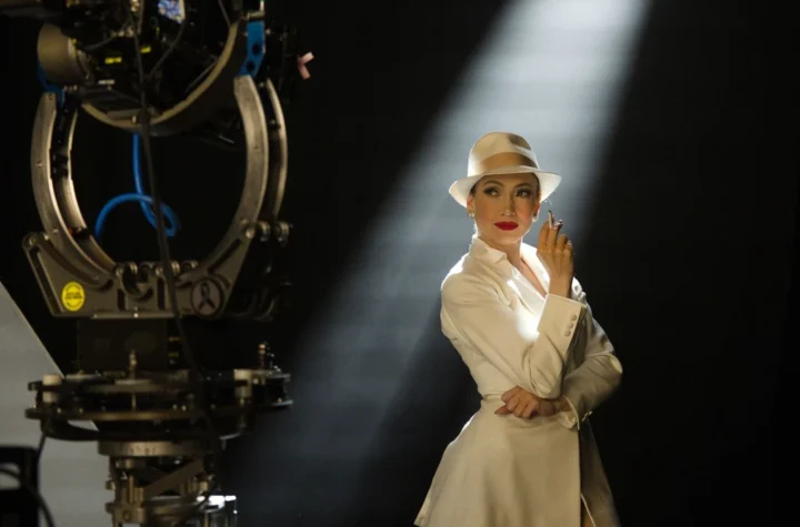 Jennifer Lopez in a white suit and fedora, lit by a shaft of light beside a vintage film camera on a soundstage, in a production still from Kiss of the Spider Woman